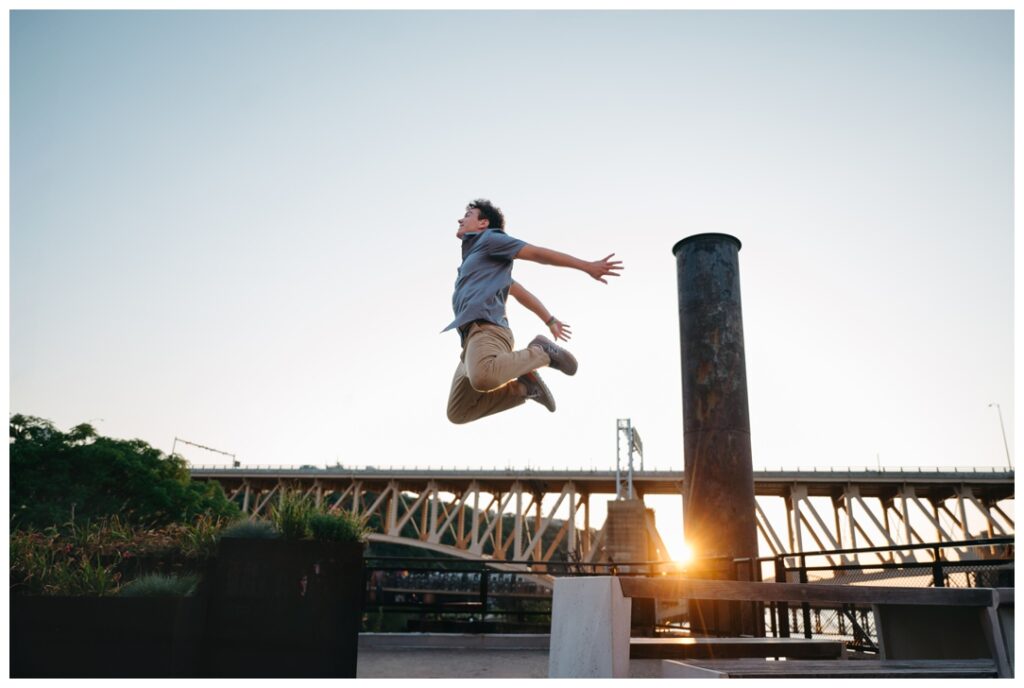 Senior Boy jumping high with a pittsburgh industrial bridge in the background at sunset 